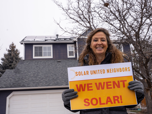Bernadette, a program participant, poses in front of her home's rooftop solar panels with a sign that reads "we went solar!"