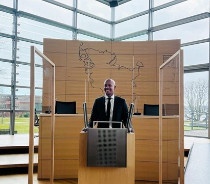 CM Zahilay at a podium, with an outline of Germany on the wall behind him