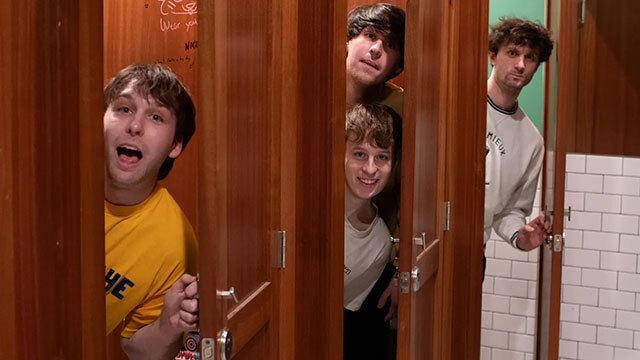 Four young men peek heads out of wooden bathroom stalls